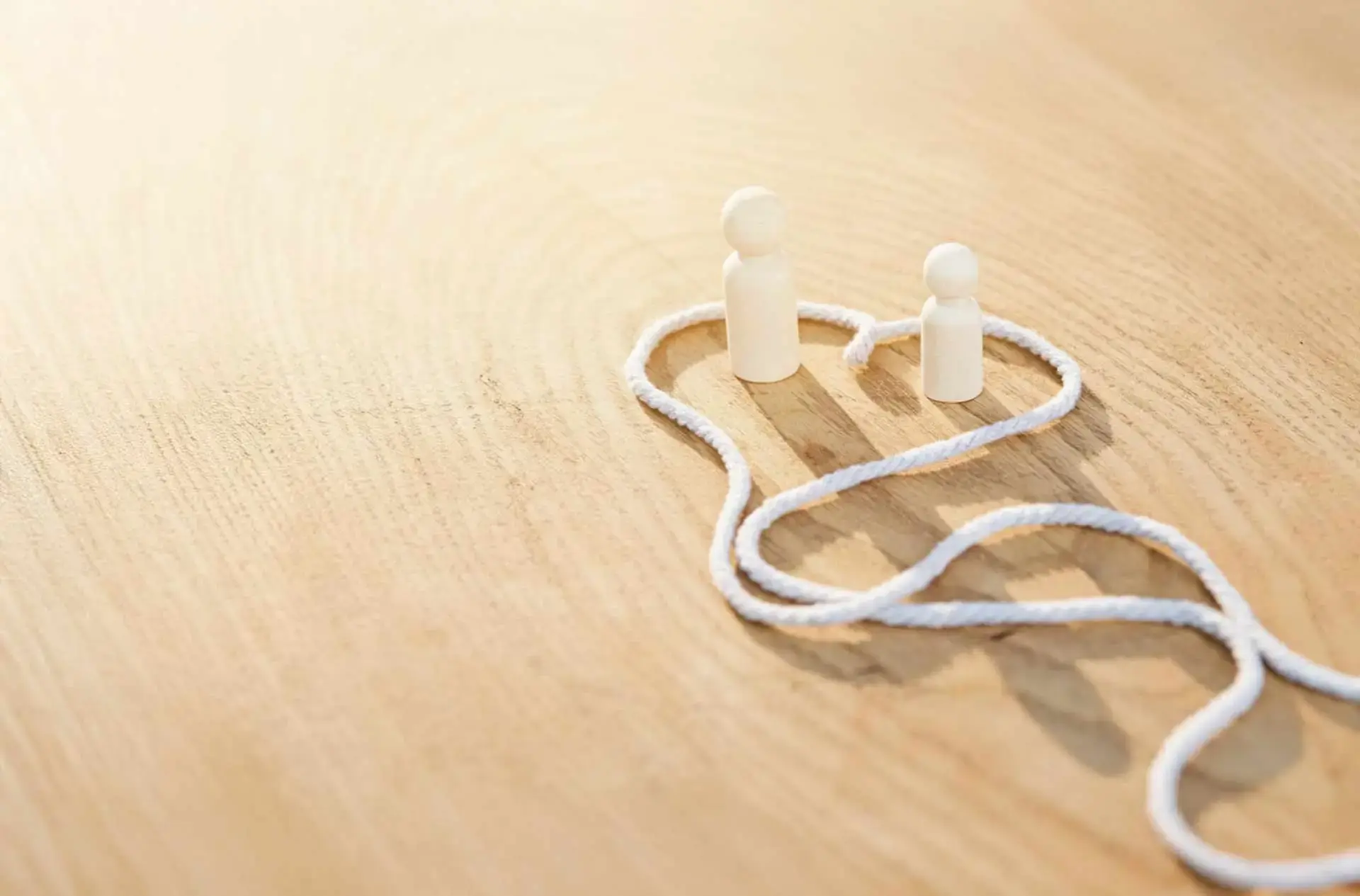 Zwei weiße Figuren stehen in einer Herzlinie aus Kordel auf hellem Holzhintergrund und symbolisieren Paarberatung, Nähe und systemische Therapie.